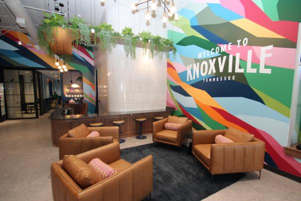 The modern interior of the Visit Knoxville Visitors Center featuring a lounge area and a vibrant mural. In the foreground, four tan leather armchairs with colorful patterned pillows are arranged around a dark area rug. The background is dominated by a large, floor-to-ceiling mural composed of bright geometric shapes in shades of blue, green, pink, and orange, with the words "WELCOME TO KNOXVILLE TENNESSEE" printed in bold white letters. To the left, a wood-paneled counter with blue barstools sits beneath a white tiled pillar and a wooden ceiling fixture overflowing with hanging green plants. Exposed bulb light fixtures hang from the ceiling, adding to the contemporary industrial feel.