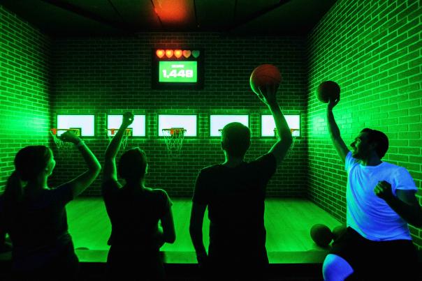 Four players shoot hoops in a neon game room at Activate Games.
