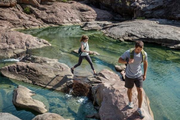 Two people hike across hill country rock to cross a clear flowing stream.