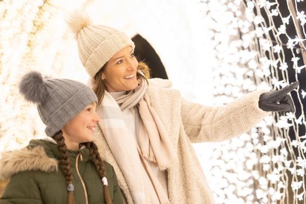 A mother and daughter wearing wooly hats, scarves and gloves in a tunnel of fairy lights