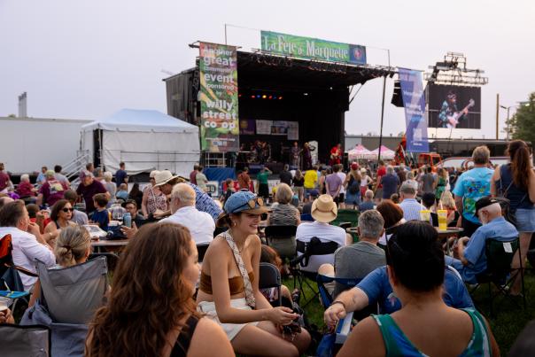 A large crowd sits and stands on a grassy area facing an outdoor concert stage at La Fête de Marquette, with musicians performing under stage lights and festival banners visible above the stage.