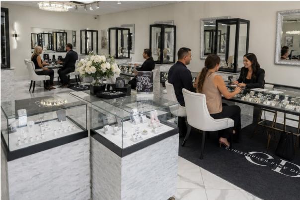 A modern, elegant, and large jewelry showroom with a couple in the background being shown jewelry by a professionally dressed sales associate at the Christopher Fine Diamonds showroom in Chandler, AZ.
