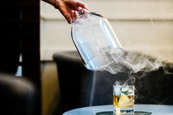 A waiter removing a glass cover that had been on a glass of whiskey to smoke it.  Smoke is still lingering in the air around the glass