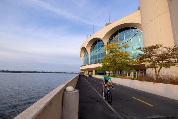 A man in a biker suit wearing a helmet as he rides his bike past the lake and Monona Terrace Convention Center.