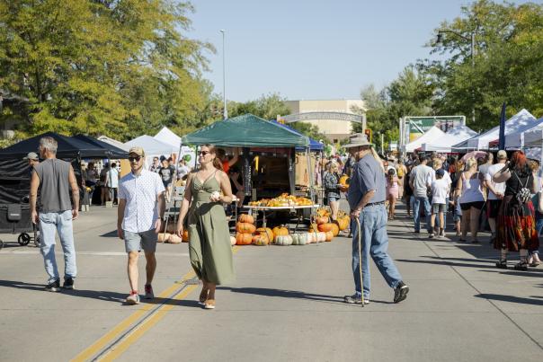 A bustling outdoor market scene with people walking along stalls under tents. Pumpkins are displayed, and trees line the sunny street, creating a festive atmosphere.
