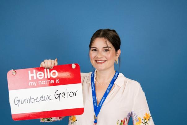 woman in floral print white blouse poses with a stuffed gator and giant name badge that says Gumbeaux Gator