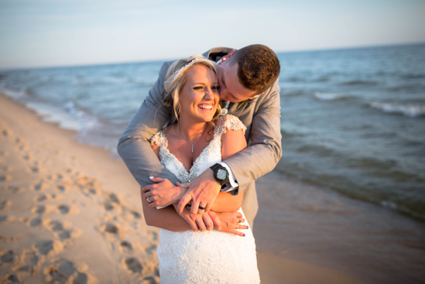 Bride and groom embrace joyfully on a sunlit beach, with gentle ocean waves in the background. The scene conveys warmth and happiness.
