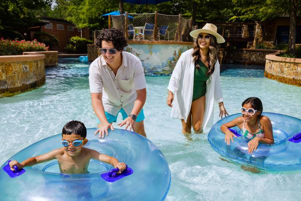 A young Latino family plays in the pool at The Woodlands Resort. Two young children (a boy and a girl) float on inner tubes while their parents gently push them along the lazy river.