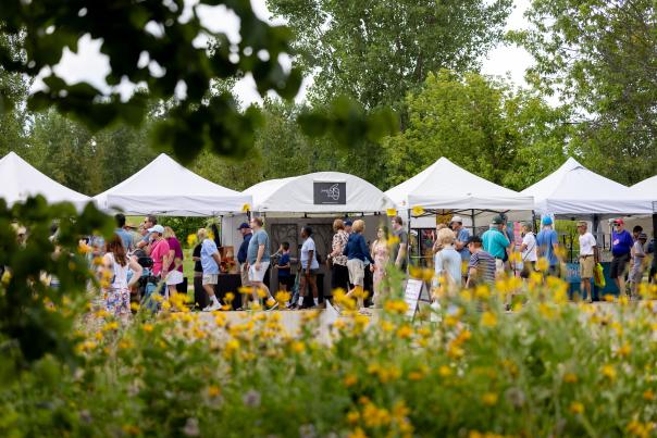 A vibrant outdoor market scene with white tents and crowds of people browsing. Colorful flowers in the foreground enhance the lively atmosphere.