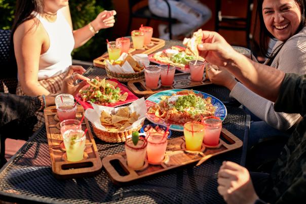 An outdoor patio with food and drinks, with people around it. Nachos, chips, enchiladas, margaritas are being enjoyed by the group of people.