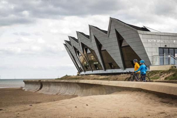 two people with bikes stood on a beach promenade in front of a sea observatory