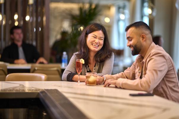 Relaxing together at the bar of Toscana Kitchen & Wine Market, a couple shares laughter and conversation in a cozy, welcoming atmosphere.