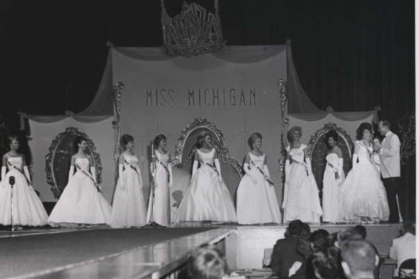 black and white photo of 9 Miss Michigan contestants on stage with interviewing man. looks like the early 1960s