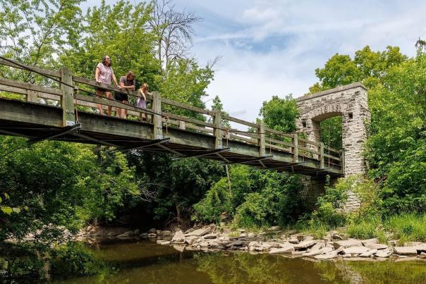 A family stands on the Hoyt Park bridge, looking over the river against a lush green background of trees