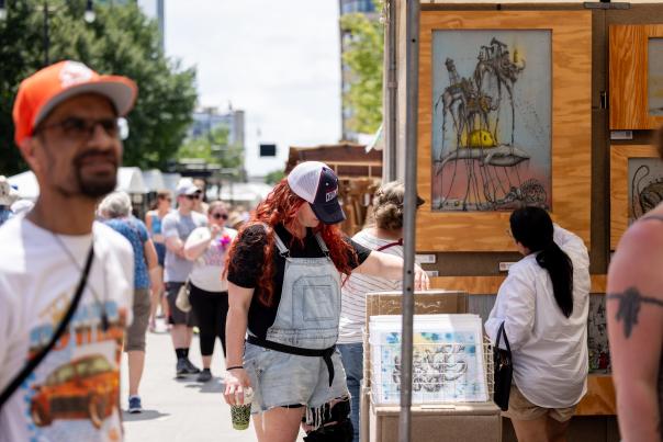 People browsing artwork at an outdoor art fair on a sunny day, with framed surreal paintings displayed in wooden booths and visitors walking past along a city street.