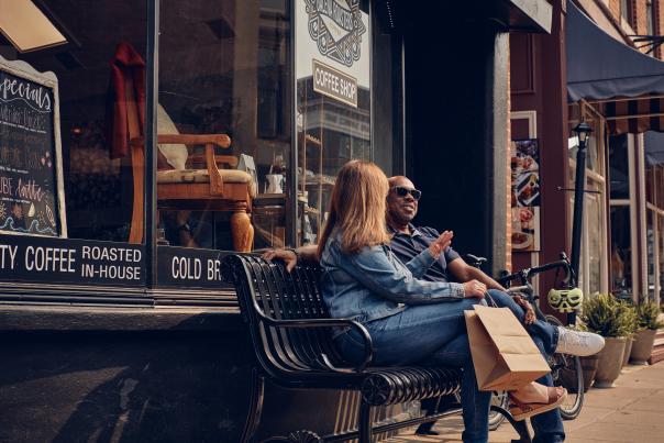 Two people sitting on a bench with a shopping bag, in front of a coffee house.