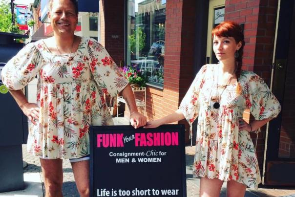 Two people wearing matching floral dresses and black boots pose beside a Funk Your Fashion consignment store sign on a sunny downtown sidewalk.