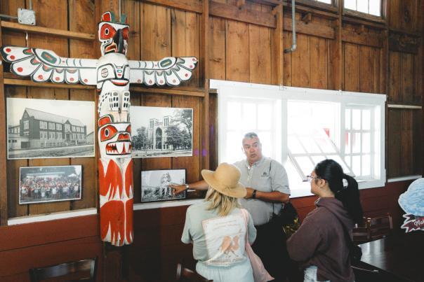 A guide explains historical photos to two visitors next to a carved totem pole inside a rustic wooden hall.
