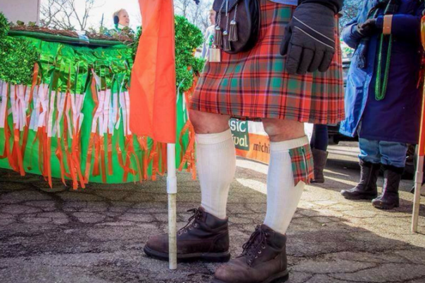 image of man from waist down holding an orange flag, wearing a traditional kilt in front of float decorated in green, white and orange with shamrocks
