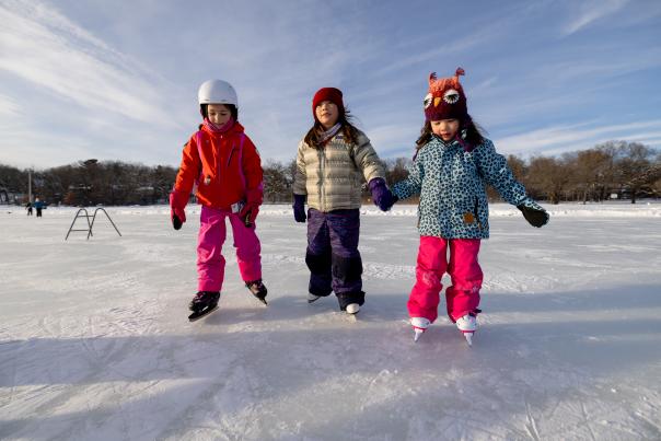 Three young girls are bundled up for winter and are holding hands while ice skating on the frozen lake.