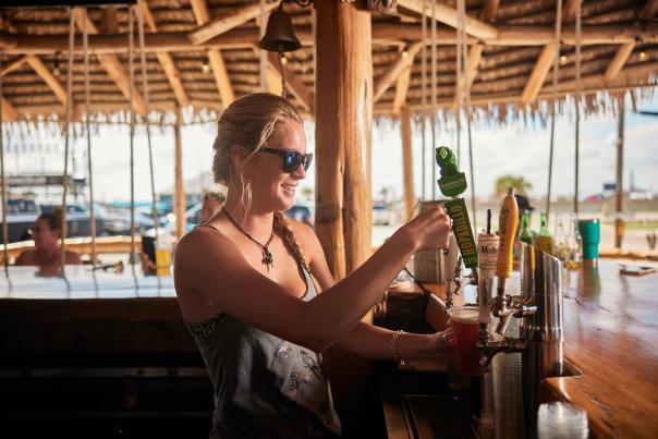 A woman under a tiki hut stands at a bar and pours a beer from tap