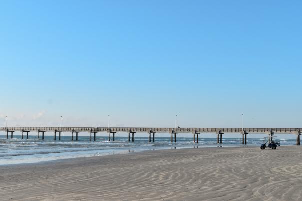 A golf cart in the distance sits on the far right of a beach image with a pier right in the middle crossing the frame.