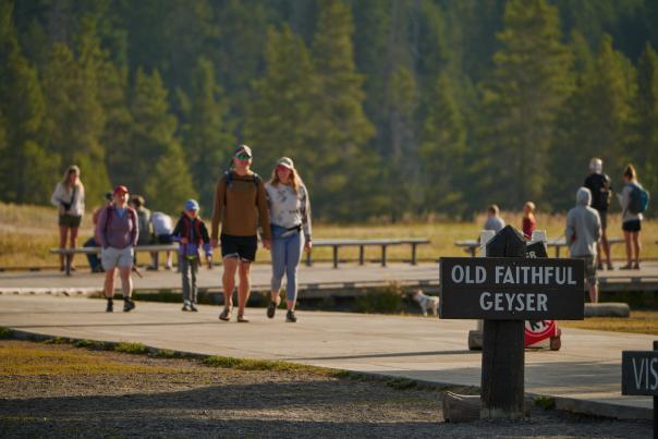 Visitors walk along the boardwalk near Old Faithful Geyser in Yellowstone National Park. A prominent Old Faithful sign marks the location, while forested landscapes and open skies provide the backdrop for one of Wyoming’s most iconic natural attractions.