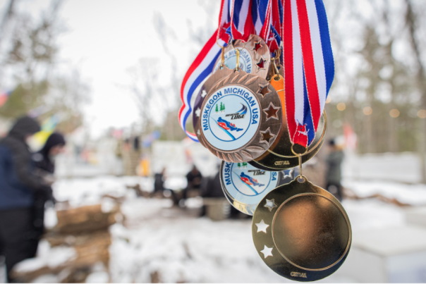 Medals with red, white, and blue ribbons hang on a snowy scene, featuring inscriptions of Muskegon, Michigan, USA. People are gathered in the background, with blurred trees and logs visible.