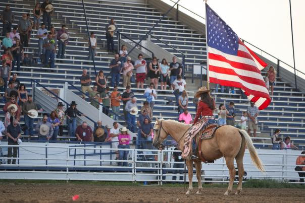 grandstands with a crowd at an area and a woman holding an American flag while on horseback