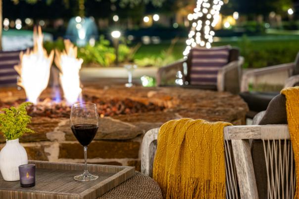 A close-up view of cushioned, light-wood chairs circled around a lit fire pit. The chair closed to the camera has a dark yellow blanket draped over one arm. A side table features a glass of wine, a small candle, and a small vase of fluffy light green plants.