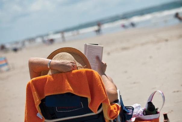 Person wearing a sunhat reads a book while reclining on a beach chair draped with an orange towel at Brunswick Islands.