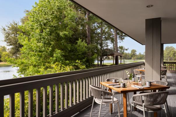 The patio at Harrison's features a row of square wood tables and gray wicker chairs beside a thick railing. A lake, bright green trees, and a gazebo lie beyond.