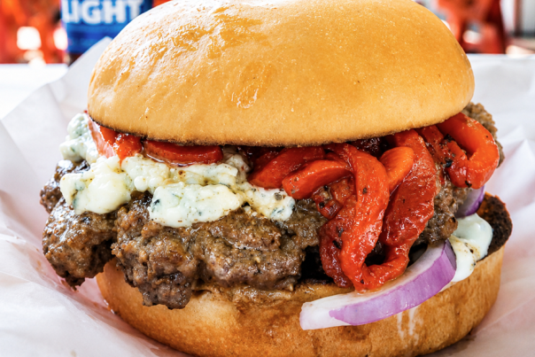 A Fourth of July Burger at Lankford's features a thick beef patty, roasted red peppers, red onion slices, ranch dressing, and blue cheese crumbles on a brioche bun. Bud Light Beer bottles stand in the background.