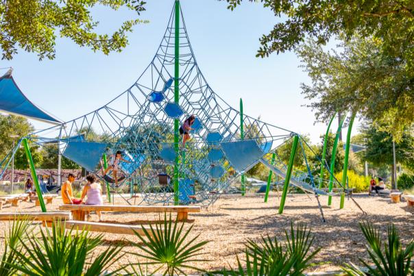 Two children climbing on a netted playscape while their parents watch.