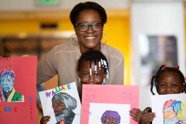 A black woman with two black children smile holding their art.