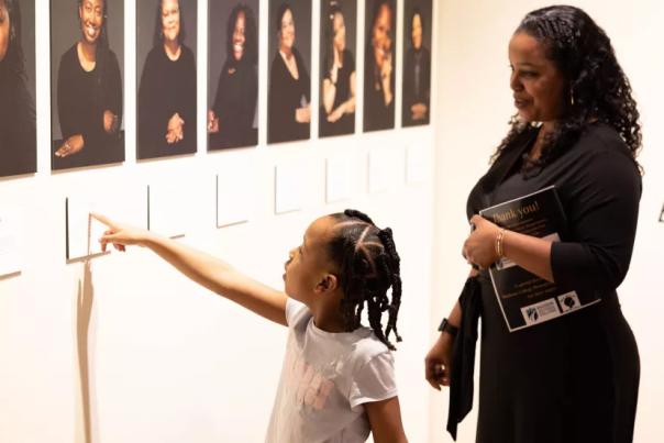 A young Black girl points at the portrait of a Black woman in an art gallery while an adult Black woman stands behind her