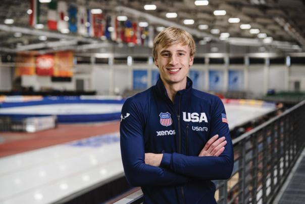 A person wearing a navy USA team jacket stands smiling on an indoor ice rink, with the rink blurred in the background. The jacket displays several sponsor logos and a small American flag patch.