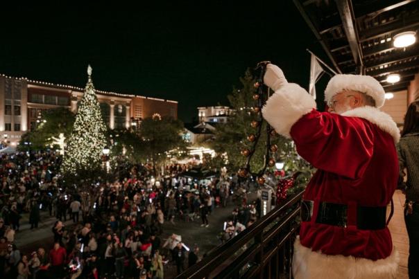 The viewer stands to the left of Santa, who waves jingle bells to a crowd from the roof of a building at Market Street, which is lit up with Christmas lights and holiday activities.