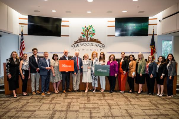 Seventeen people line up in front of dais in The Woodlands Township Board Chambers. They are all Visit The Woodlands Board Members and Staff. From left to right: Josie Lewis (Director of Sales), Brooklyn Newell (Social Media Coordinator), Ryan Greenwood (Senior Sales Associate), Nick Wolda (President), Cameron Klepac (Secretary / Treasurer of the Board), Brad Bailey (Director of the Board), Richard Franks (Vice Chairman of the Board), Linda Nelson (Chairman of the Board), Kim Lowe (Director of the Board), Noemi Gonzalez (Director of the Board), Monique Sharp (Director of the Board), Elizabeth Eddins (Executive Director), Carla Twiddel (Administrative Analyst), Julie Quinn (Servicing Specialist), Amber Parsley (Tourism Specialist), Ashley White (Director of Marketing), Andrea Day (Creative Media Coordinator). Missing: Ashley Fenner (Senior Sales Associate) and Rachael Varner (Content Coordinator)