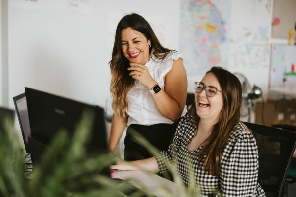 Two people laughing, one sat at a desk, the other stood over their shoulder.