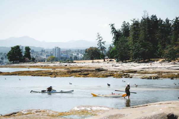 One person paddles a kayak while another walks beside a kayak in shallow water along a rocky shoreline.