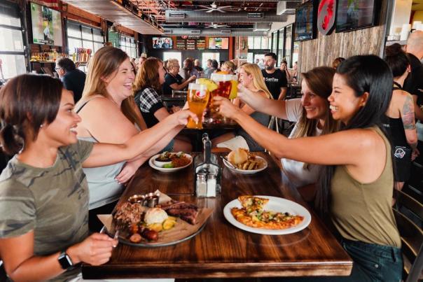 a busy restaurant interior in the background with focus on a table filled with plates of food and four ladies raising a glass of beer to cheer with