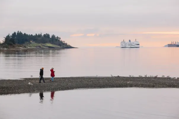 Two people walking on a little bit of land with a ferry in the background.