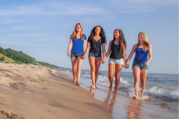 four teen girls hold hands while walking along shoreline of Lake Michigan