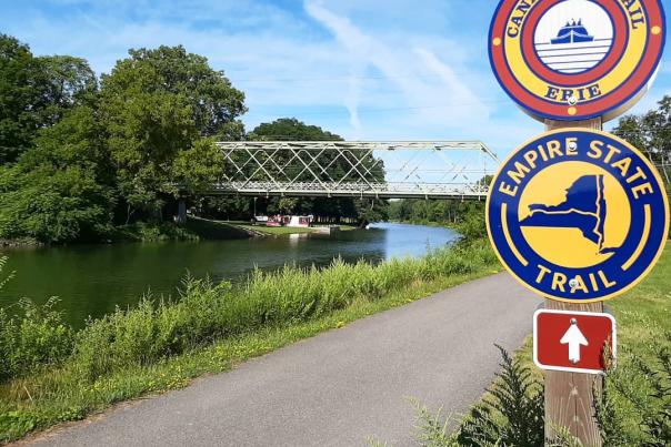 In the foreground a wooden sign post stands with two circular signs stacked vertically. The top sign reads "Canalway Trail" on the top half of the circle and "Erie" on the bottom half, while the bottom sign reads "Empire State" on the top half of the circle and "Trail" at the bottom.

In the background sits the Erie Canal in Brockport, NY.