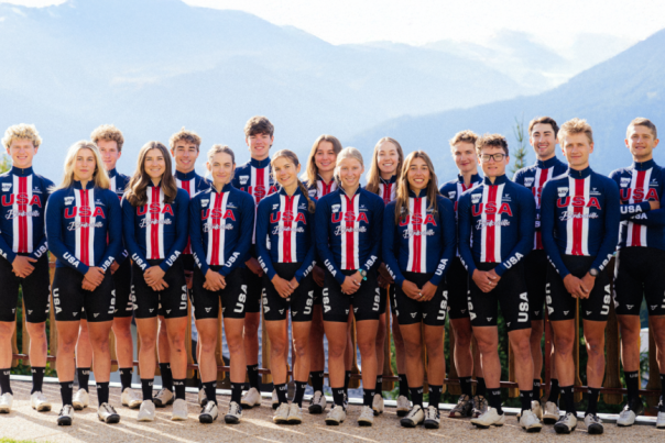 Sixteen USA cycling team members in matching blue, red, and white uniforms pose on a wooden deck with mountains in the background