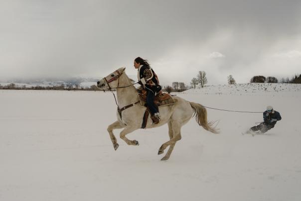 A horseback rider pulls a skier across a snow-covered field during a skijoring run near Jackson, Wyoming. The horse gallops through deep snow as the skier follows behind, highlighting the combination of Western horse culture and winter recreation in Teton County.