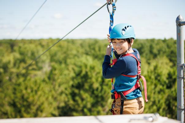 Young boy smiling and holding onto his harness at the top of the zip line platform at NEW Zoo & Adventure Park.