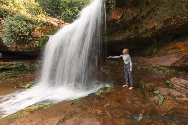 A young woman stands barefoot on a reddish-brown rock ledge, reaching toward the powerful, white curtain of a waterfall that flows over a mossy, rocky cliff face.