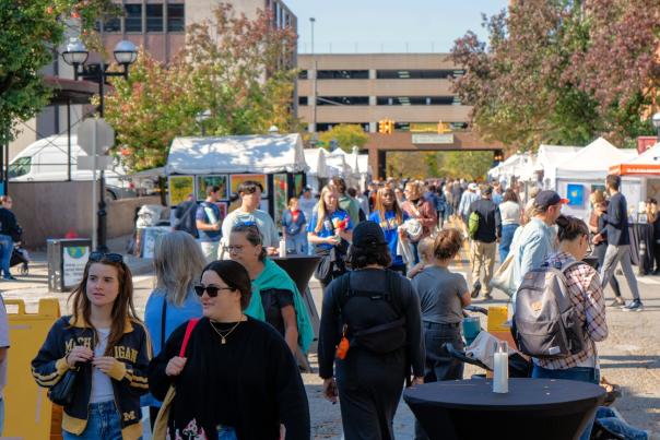 People walk around a closed street in Ann Arbor browsing white vendor booths line the street filled with art. Fall time of year with orange and brown leaves on the trees in the background.
